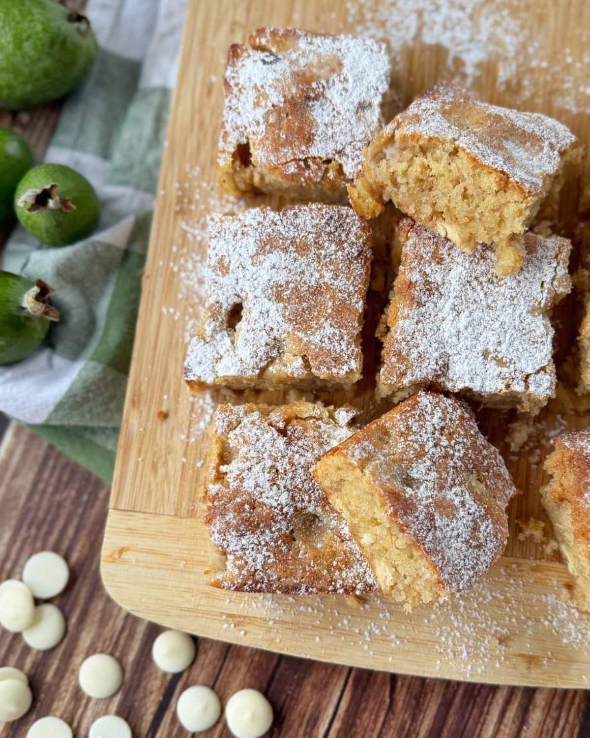 Feijoa and White Blondie Several pieces of feijoa blondie dusted with powdered sugar are arranged on a wooden board, with white chocolate chips and green fruits nearby.