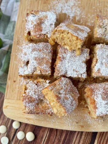 Nine pieces of powdered sugar-dusted blondies are arranged on a wooden board, with feijoas, a striped napkin, and white chocolate buttons nearby.