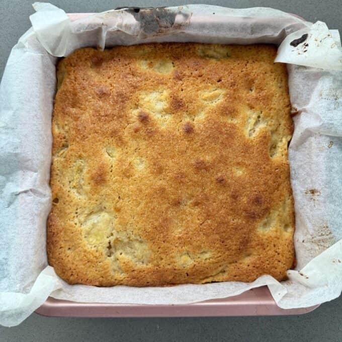 Feijoa and White Blondie Golden brown square cake in a parchment-lined baking pan, with a slightly uneven surface and visible baked spots.