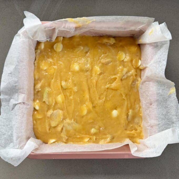 Feijoa and White Blondie Square baking pan lined with parchment paper filled with unbaked yellow batter containing visible white chocolate chips and banana pieces.
