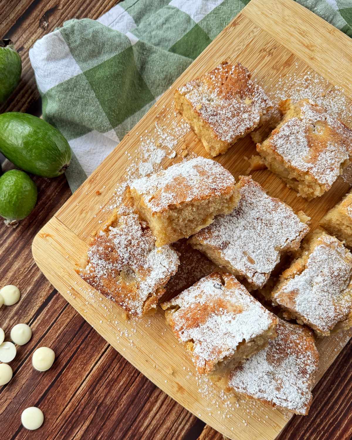 Feijoa and White Blondie Nine powdered sugar-dusted feijoa blondie squares are arranged on a wooden board, with whole green feijoas, white chocolate chips, and a green checkered cloth nearby.