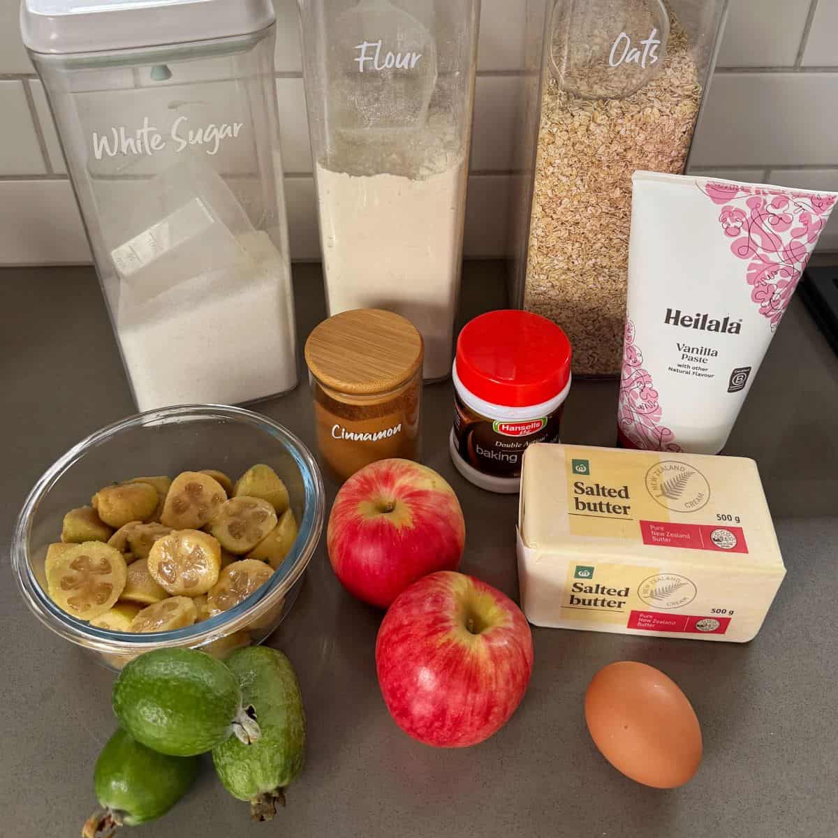 Feijoa and Apple Crumble Slice A countertop displays white sugar, flour, oats, cinnamon, baking powder, vanilla, two apples, feijoas, sliced kiwifruit, two blocks of salted butter, and an egg.
