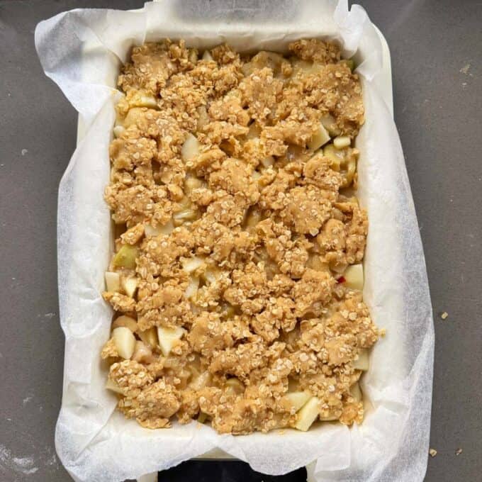 Feijoa and Apple Crumble Slice A rectangular baking dish lined with parchment paper, filled with diced apples and topped with an oat crumble mixture, ready to be baked.