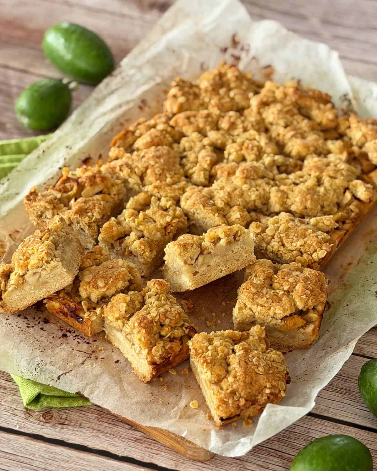 Feijoa and Apple Crumble Slice A tray of crumbly, golden-brown dessert bars cut into squares sits on parchment paper, with green feijoa fruits placed around it on a wooden surface.