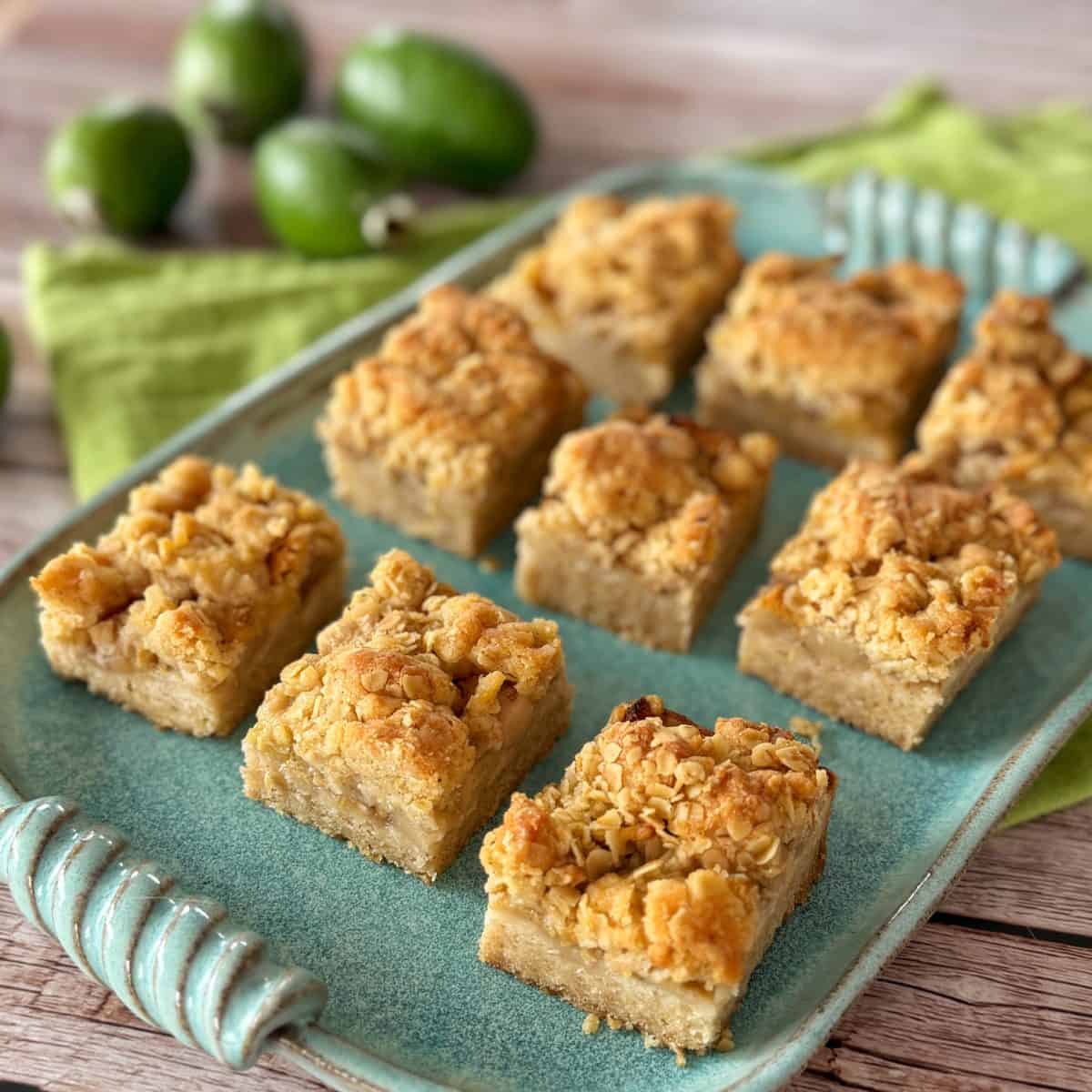 Feijoa and Apple Crumble Slice A rectangular blue plate holds nine pieces of crumbly, golden-brown dessert bars arranged in three rows; green fruits and a green cloth are in the background.