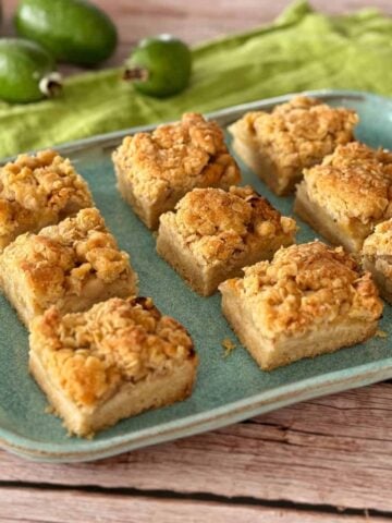 A blue ceramic plate with nine square crumb-topped dessert bars on a wooden surface, with green feijoas and a green cloth in the background.