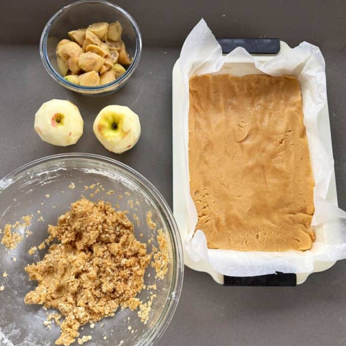 Feijoa and Apple Crumble Slice Baking ingredients prepared: a baking tray lined with parchment and dough, two peeled apples, chopped apples in a bowl, and oat mixture in a glass bowl on a countertop.