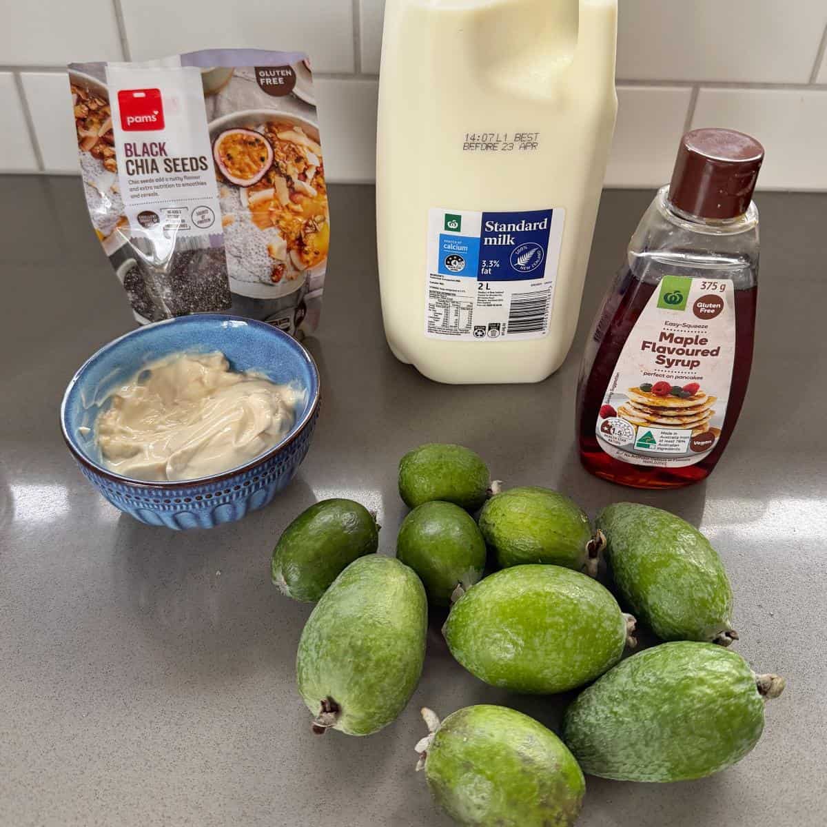 A bowl of yogurt, a packet of black chia seeds, a bottle of milk, maple flavoured syrup, and several fresh feijoas on a kitchen counter.