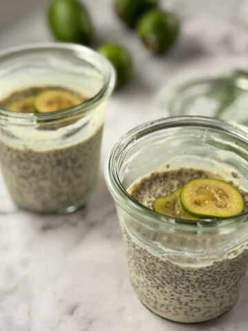 Two glass jars filled with chia seed pudding, each topped with slices of green fruit, sit on a marble surface. Blurred green fruits are in the background.