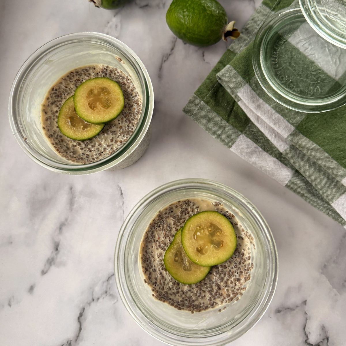 Feijoa Chia Puddings Two glass jars of chia pudding topped with two slices of feijoa each, placed on a marble surface near a green and white cloth and whole feijoas.