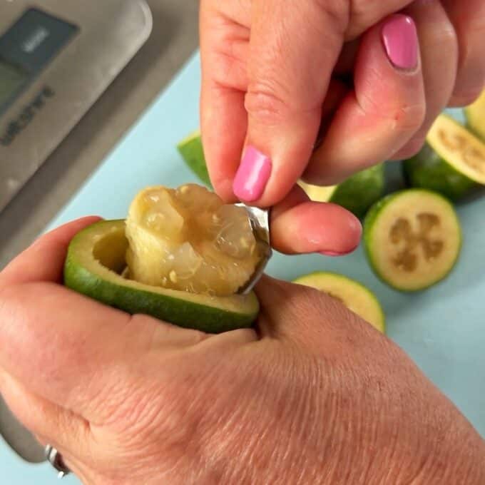 Feijoa Chia Puddings A person scoops out the pulp from a halved feijoa fruit using a spoon, with several cut feijoas visible on a cutting board.