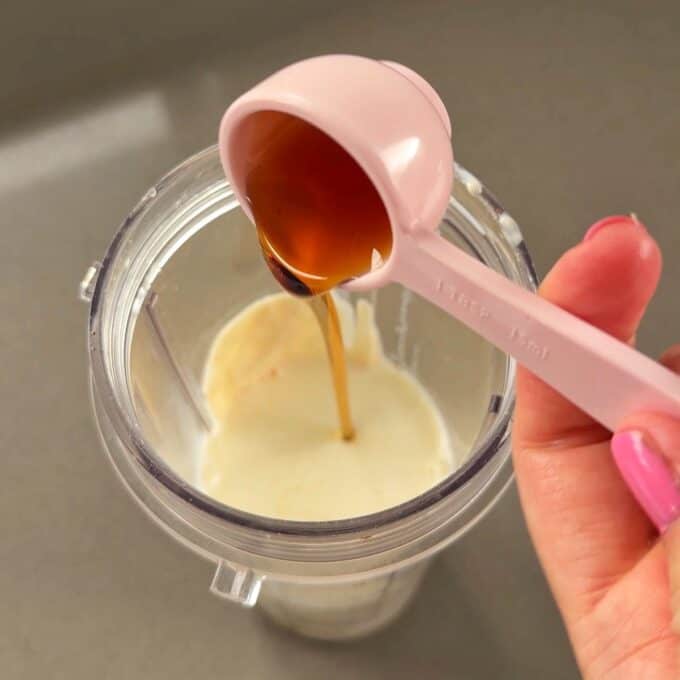 Feijoa Chia Puddings A hand holds a pink measuring spoon pouring liquid sweetener into a blender cup filled with milk.