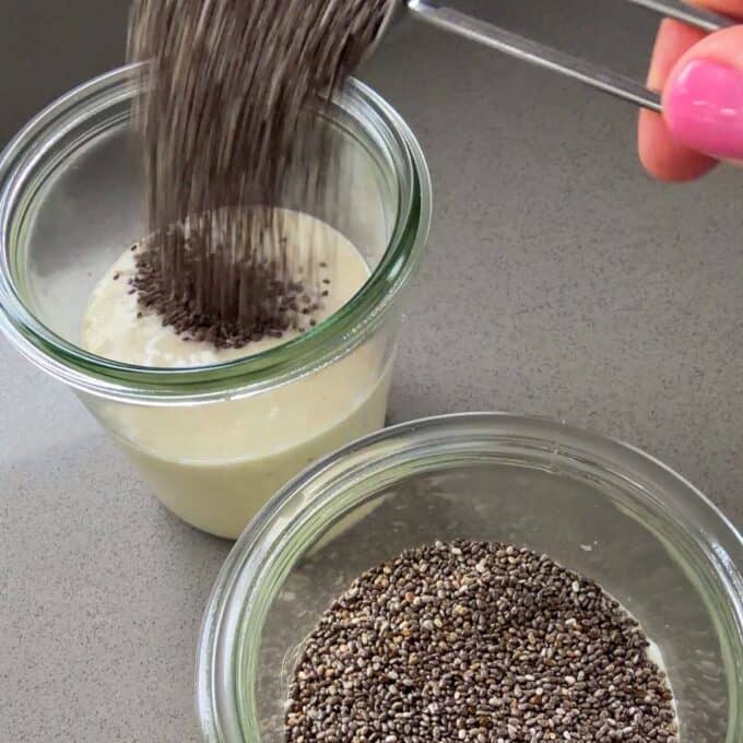 Feijoa Chia Puddings A hand pours chia seeds from a metal scoop into a glass jar containing a creamy liquid, with another jar of chia seeds nearby on a gray countertop.