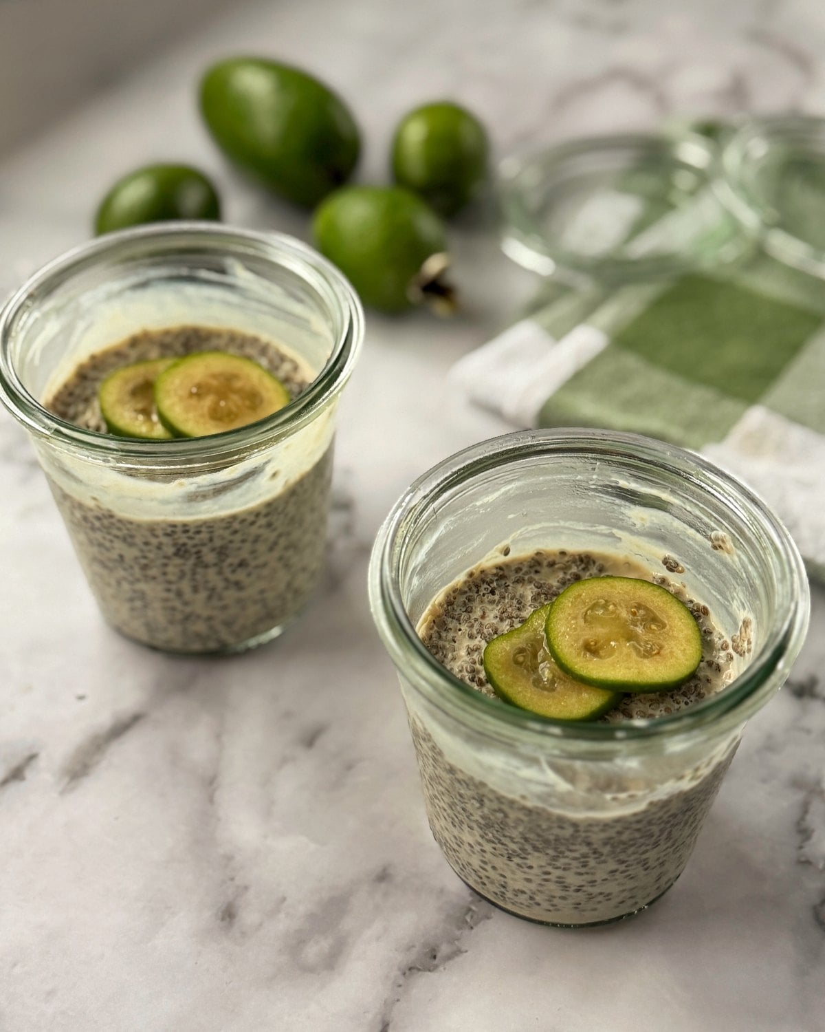 Two glass jars filled with chia pudding, each topped with two slices of green fruit, sit on a marble surface near whole feijoas and a green checked cloth.