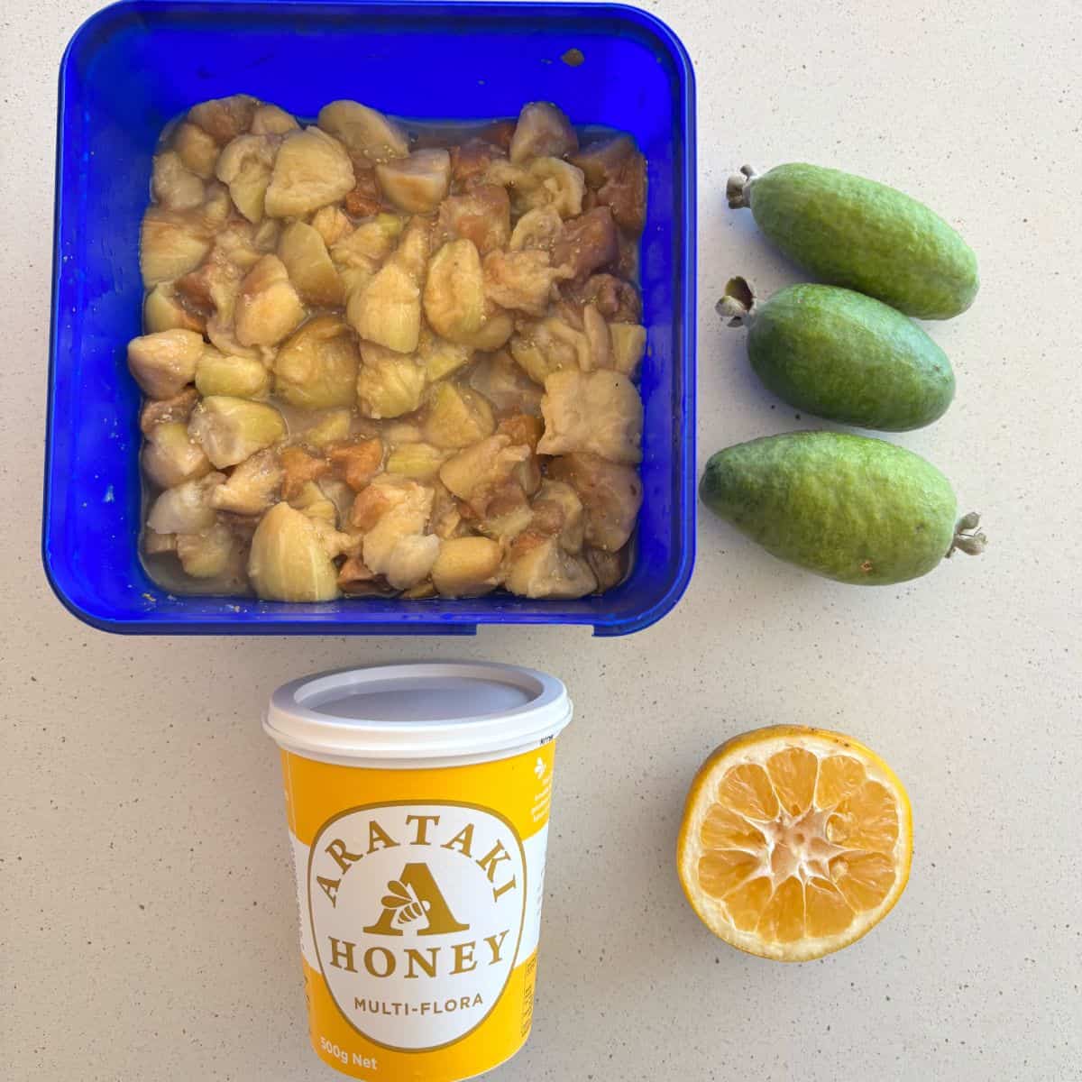 Chopped fruit in a blue container, three feijoas, a halved lemon, and a tub of Arataki multi-flora honey on a beige surface.