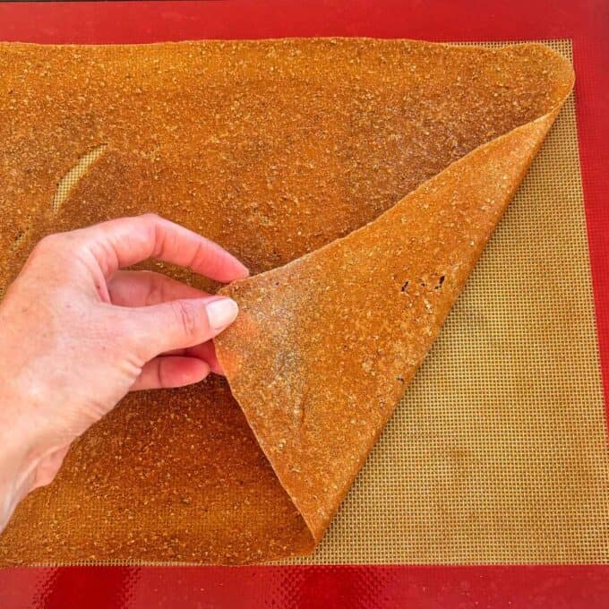 Feijoa Leather A hand lifts a sheet of homemade fruit leather from a silicone baking mat on a red surface.