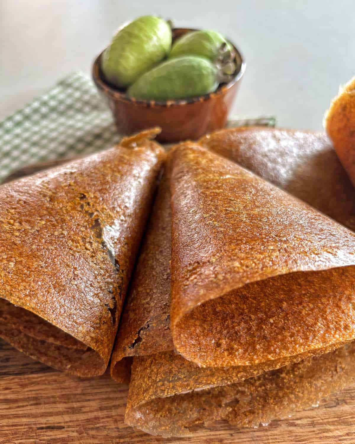 Close-up of several rolled fruit leather pieces on a wooden surface, with a small bowl of green fruits in the background.