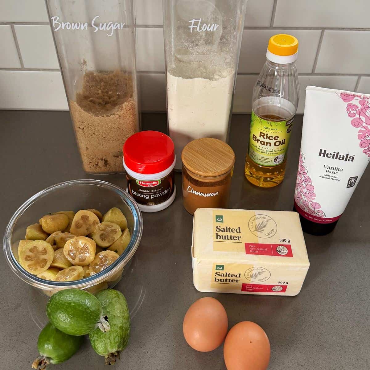 Feijoa Loaf Baking ingredients on a counter: brown sugar, flour, baking powder, cinnamon, rice bran oil, vanilla, butter, eggs, and a bowl of sliced feijoas.