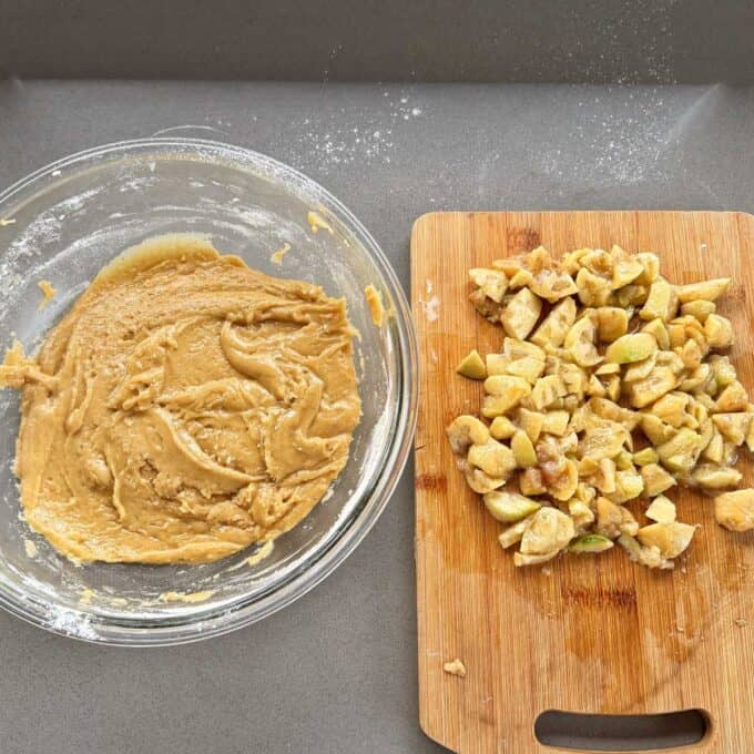Feijoa Loaf A glass bowl with thick cookie dough sits next to a wooden cutting board covered with chopped apples on a grey countertop.
