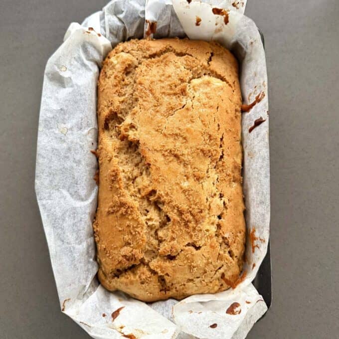 Feijoa Loaf A loaf of baked bread with a cracked top sits in a parchment-lined rectangular baking tin on a gray surface.