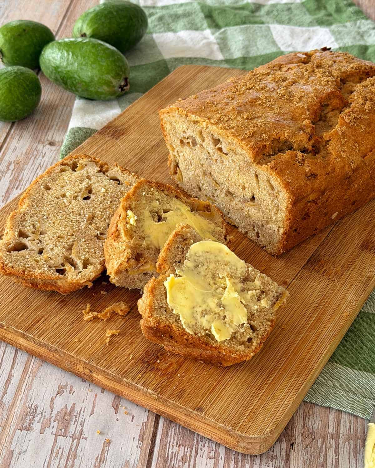 Feijoa Loaf A loaf of sliced bread sits on a wooden cutting board, with two slices buttered; green feijoa fruits and a green-checked cloth are nearby.