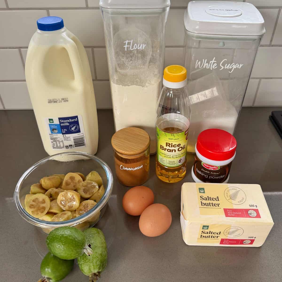 Feijoa Muffins Ingredients including milk, flour, sugar, rice bran oil, cocoa powder, cinnamon, eggs, butter, sliced bananas, and feijoas arranged on a kitchen counter.