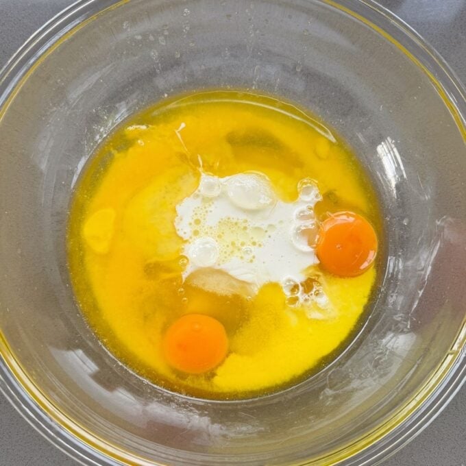 Feijoa Muffins A clear glass bowl with two raw eggs, milk, and melted butter on a gray countertop, ready to be mixed.