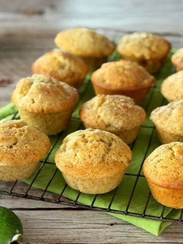 Twelve golden-brown muffins cooling on a wire rack lined with a green cloth, with a few green fruits placed nearby on a wooden surface.