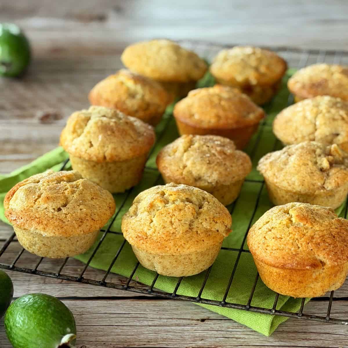 Feijoa Muffins Twelve golden-brown muffins cooling on a wire rack lined with a green cloth, with a few green fruits placed nearby on a wooden surface.