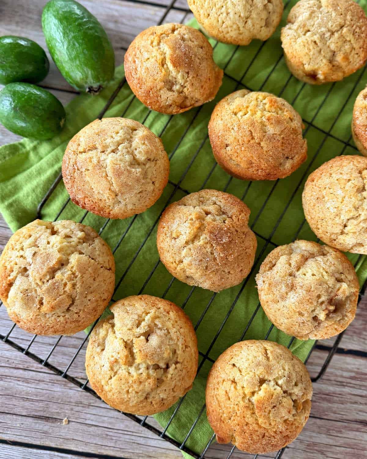 Freshly baked muffins cooling on a wire rack with three feijoa fruits placed nearby on a green cloth, all set on a wooden surface.