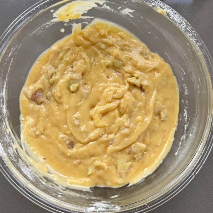 Feijoa Muffins A glass bowl containing a thick, yellowish batter with visible chunks, likely banana bread or cake mix, on a gray countertop.