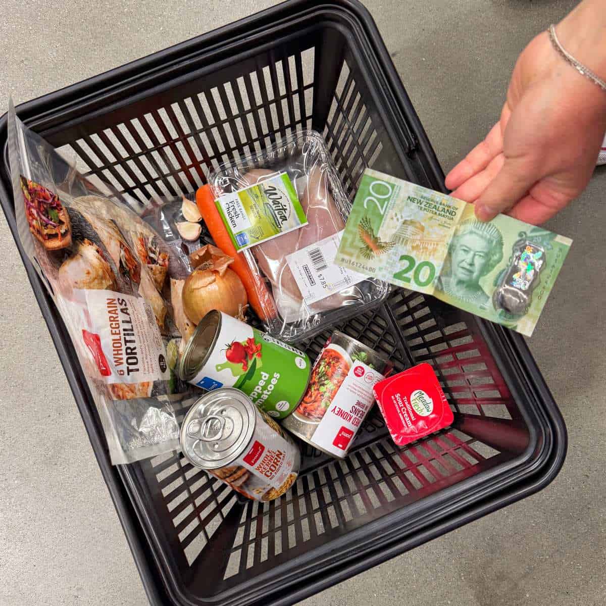 Loaded Chicken Tortilla Cups A shopping basket with tortillas, canned goods, vegetables, chicken, and a hand holding a Canadian twenty-dollar bill and coins above it.