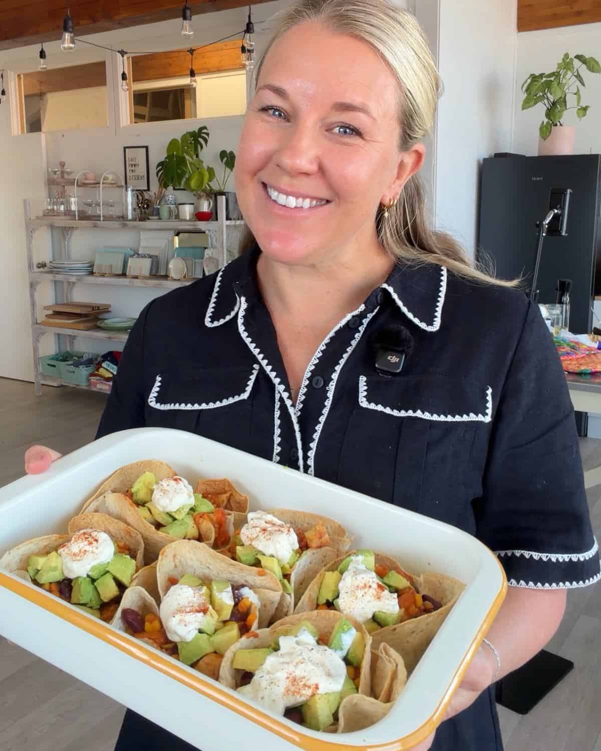Loaded Chicken Tortilla Cups A woman in a black dress holds a tray of tacos topped with avocado and sour cream in a kitchen setting.