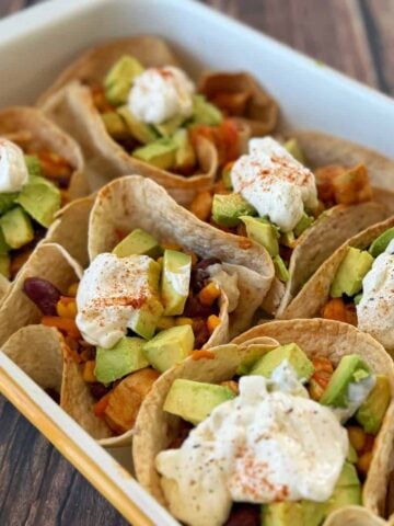 Soft tortillas filled with mixed vegetables, beans, corn, avocado chunks, and topped with a dollop of sour cream and sprinkled with paprika, arranged in a white dish.