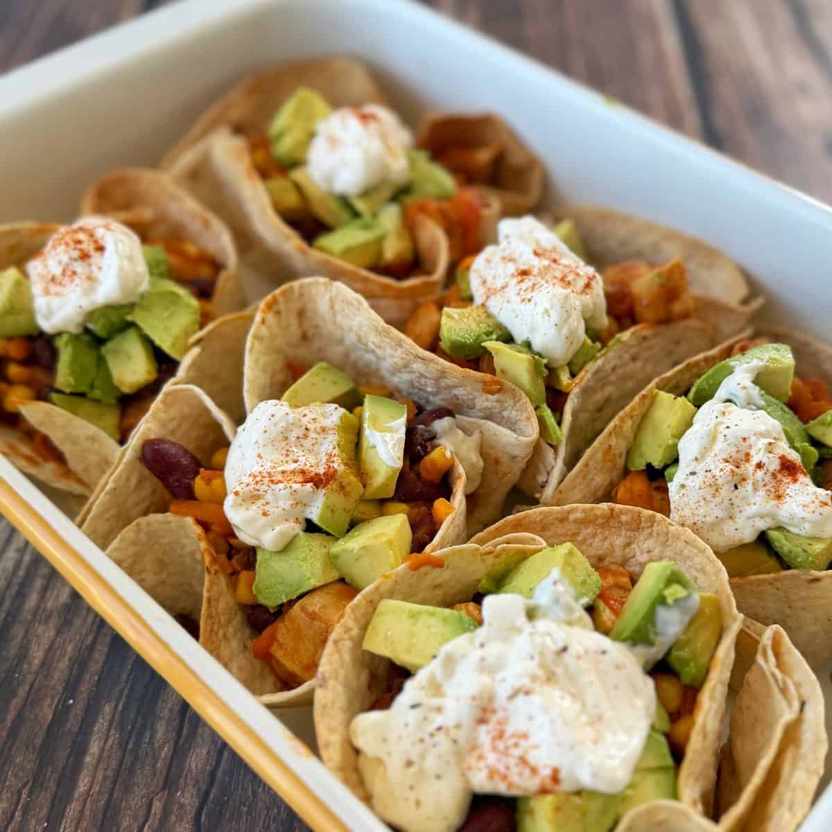 Soft tortillas filled with mixed vegetables, beans, corn, avocado chunks, and topped with a dollop of sour cream and sprinkled with paprika, arranged in a white dish.