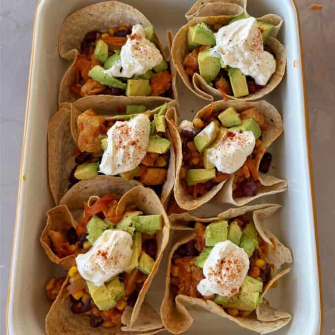 Loaded Chicken Tortilla Cups Six tortilla cups filled with a mixture of beans, corn, and vegetables, topped with diced avocado, a dollop of sour cream, and a sprinkle of paprika, arranged in a baking dish.