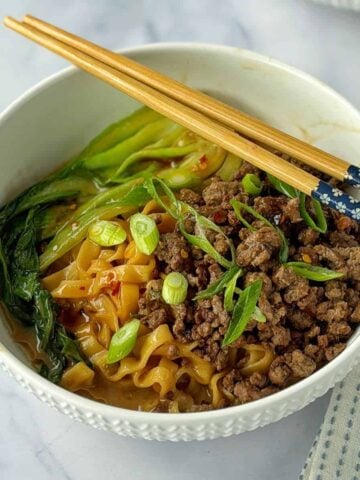A bowl of noodles topped with seasoned ground meat, sliced green onions, leafy greens, and chopsticks resting on the rim.