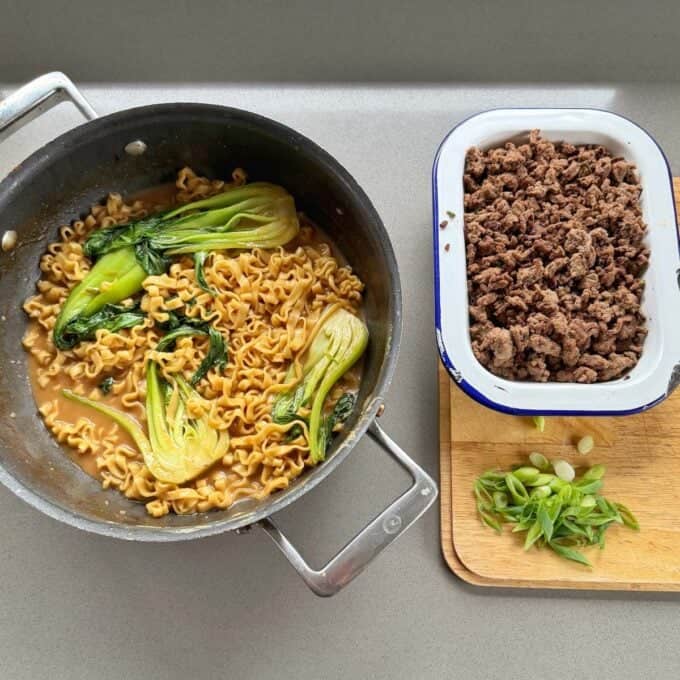 One Pot Beef Mince Noodles A pot of cooked noodles with bok choy, a tray of cooked ground beef, and sliced green onions on a cutting board are arranged on a countertop.