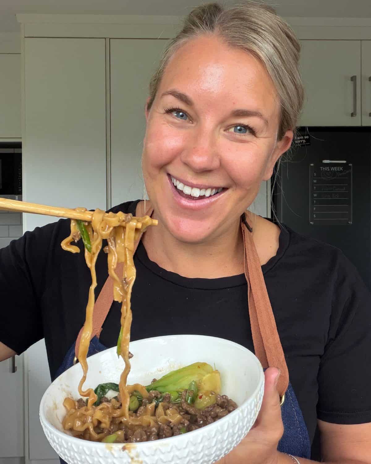 One Pot Beef Mince Noodles Woman smiling and holding a bowl of noodles with vegetables, using chopsticks to lift some noodles, in a kitchen setting.