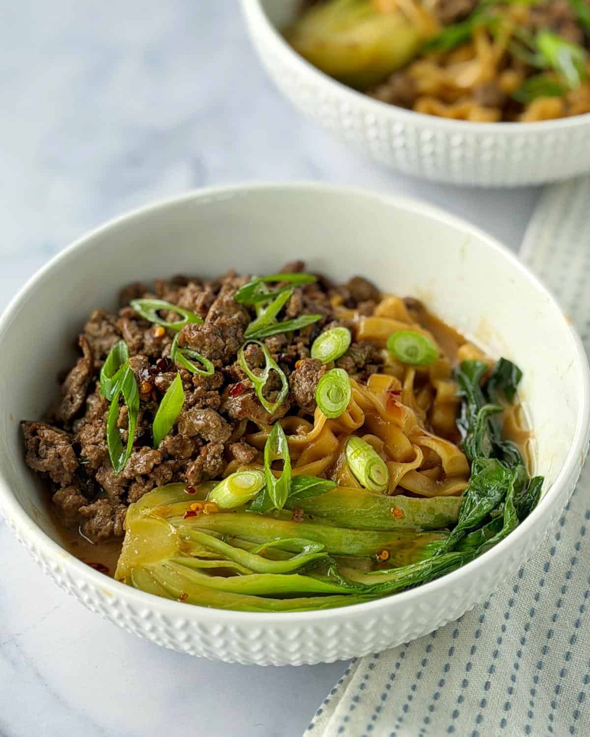 One Pot Beef Mince Noodles A white bowl filled with ground beef, wide noodles, bok choy, and sliced green onions, placed on a light surface next to a blue-striped cloth.