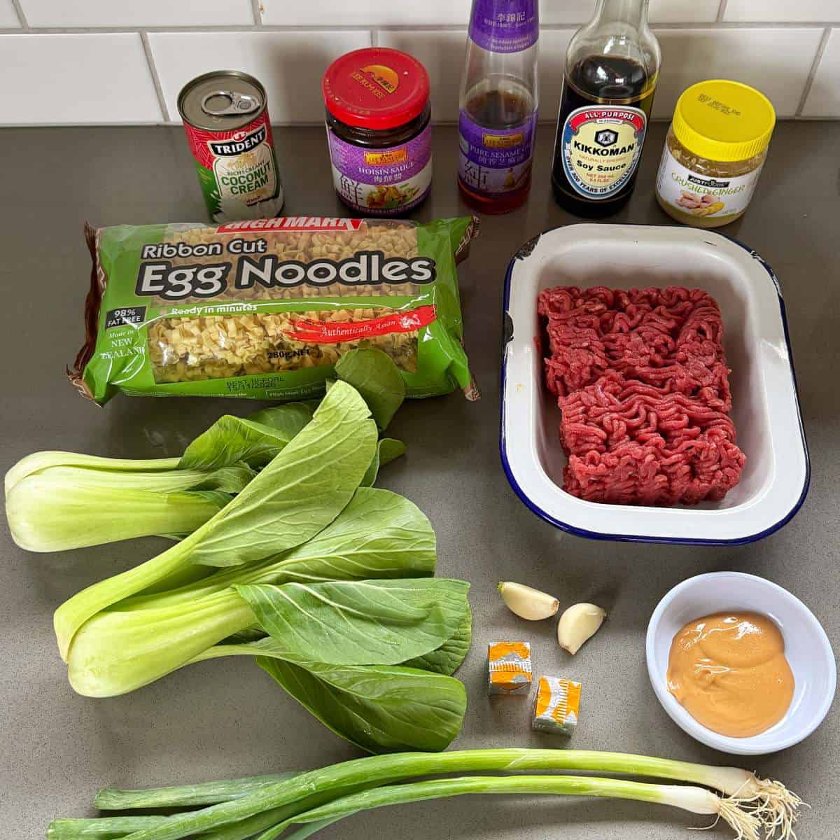 One Pot Beef Mince Noodles Ingredients for a noodle dish on a countertop, including egg noodles, ground meat, bok choy, green onions, garlic, seasonings, peanut butter, and various sauces.
