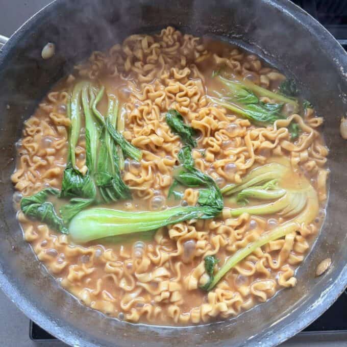 One Pot Beef Mince Noodles A pan of cooked noodles in a brown broth with baby bok choy, shown simmering on a stovetop.