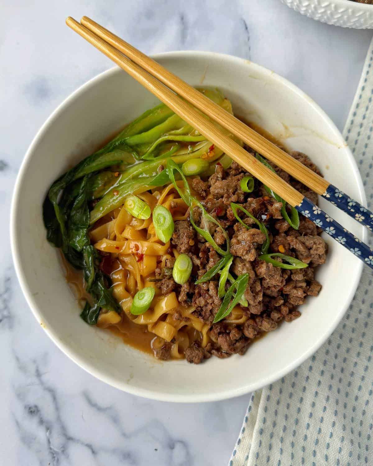 One Pot Beef Mince Noodles A bowl of noodles topped with ground meat, sliced green onions, leafy greens, and red chili flakes, with a pair of chopsticks resting on the bowl.