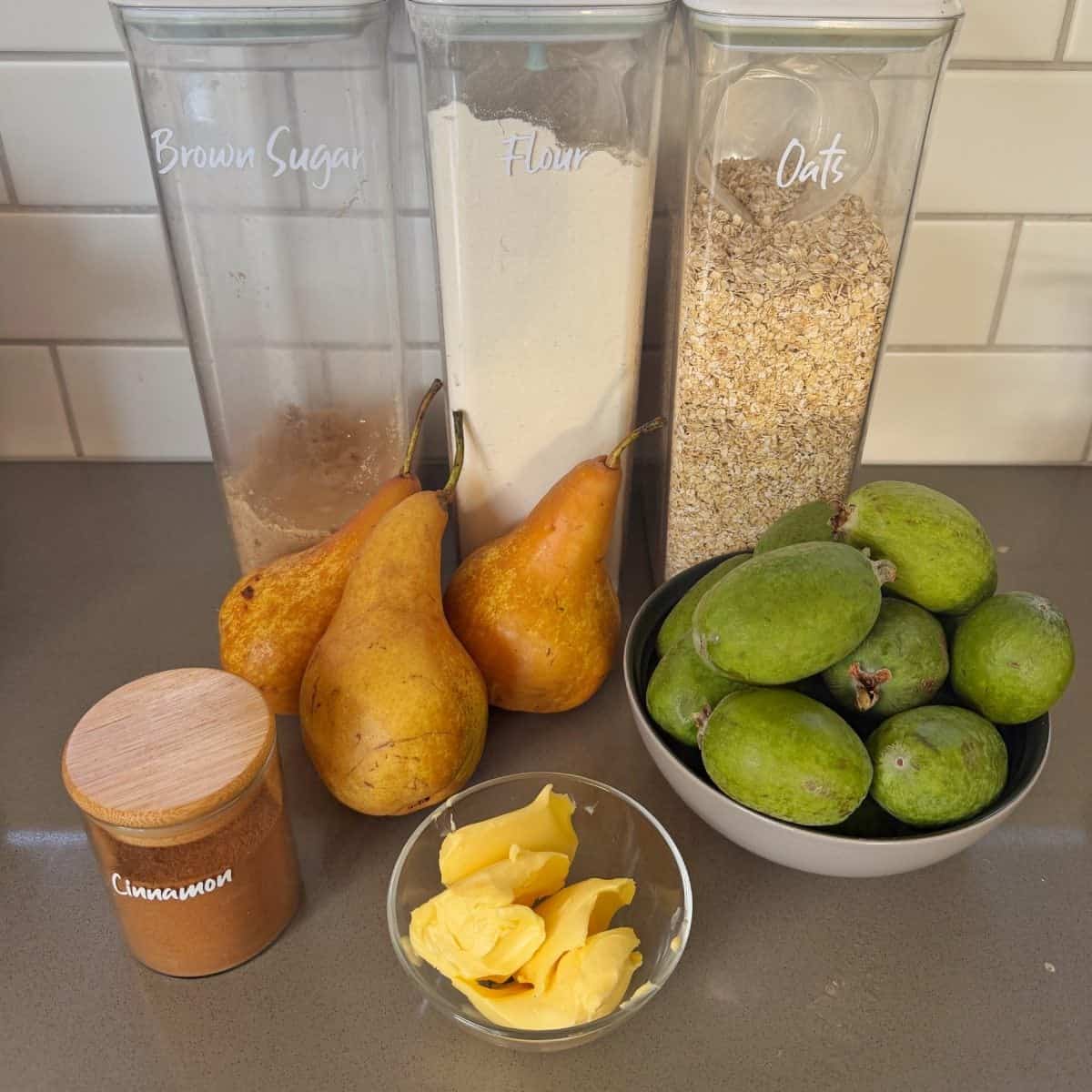 Ingredients on a countertop: brown sugar, flour, oats, cinnamon, three pears, a bowl of feijoas, and a small dish of butter.
