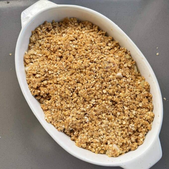 Feijoa and Pear Crumble An oval white baking dish filled with unbaked apple crumble topped with an oat and crumble mixture sits on a gray countertop.
