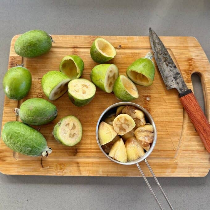Feijoa and Pear Crumble A wooden cutting board with whole and halved feijoas, a knife with a wooden handle, and a metal strainer holding scooped feijoa pulp.
