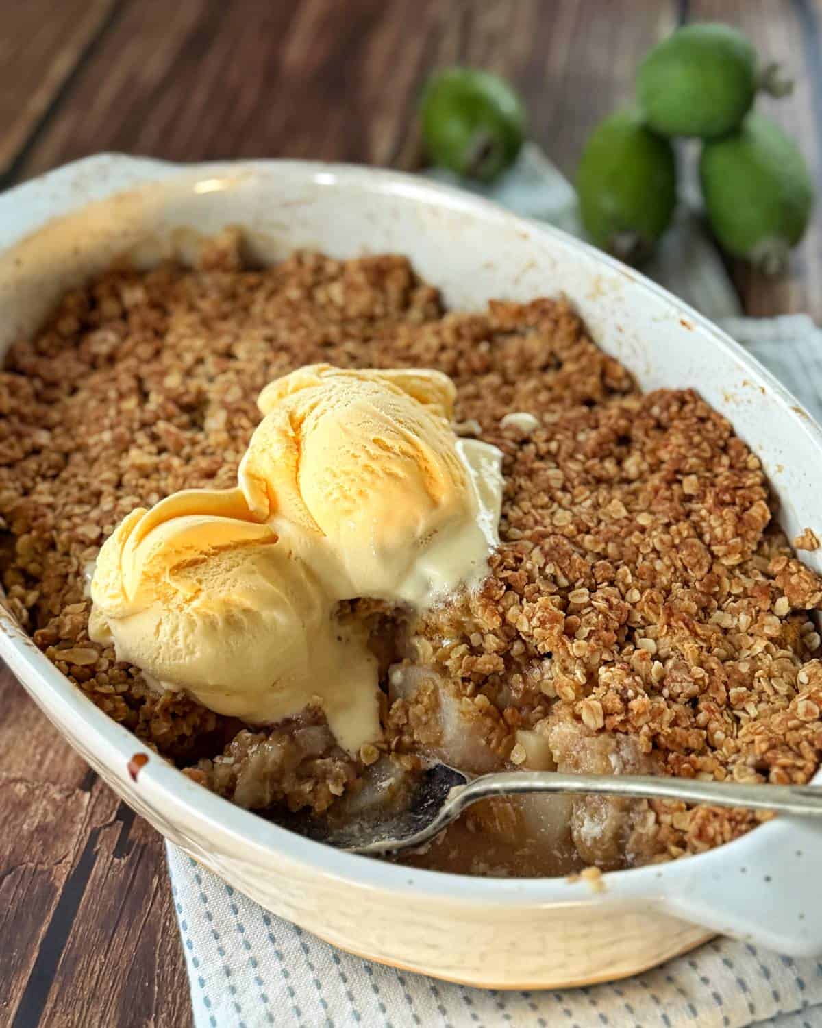 A baked fruit crumble topped with two scoops of vanilla ice cream in a white dish, with a serving spoon and green fruits in the background.