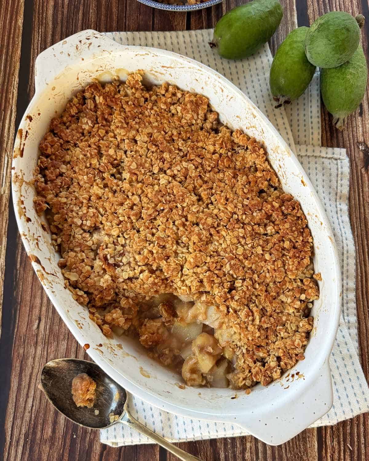 Feijoa and Pear Crumble Baked fruit crumble with oat topping in a white dish, partially served with a silver spoon; feijoas and a cloth napkin are nearby on a wooden surface.