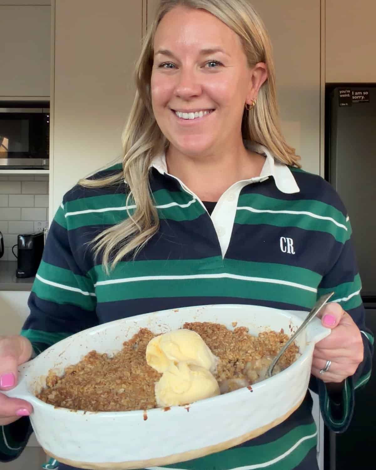 Feijoa and Pear Crumble Woman in a striped shirt holding a baking dish filled with crumble dessert topped with two scoops of vanilla ice cream.