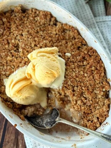 A baked fruit crumble topped with scoops of vanilla ice cream in a white dish with a spoon, placed on a light cloth on a wooden surface.